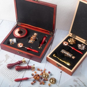 May include: Two wooden boxes containing wax seal kits with metal melting spoons, red wooden handles, glass vials of wax beads, and brass stamp heads. Additional loose wax beads and three wax seal stamps with red handles are arranged on the surface.