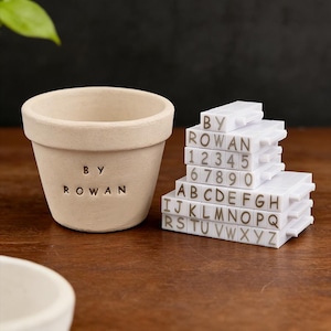 Pode incluir: Um vaso de flores de cerâmica bege com o texto "BY ROWAN" e uma pilha de carimbos brancos de letras e números. Os carimbos incluem as letras A-Z e os números 1-0. O vaso está sobre uma superfície de madeira.