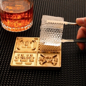 May include: A gold-coloured ice mould with four different designs, including a honeycomb pattern. A clear ice cube is held by tongs above the mould. A glass of amber liquid sits in the background. The mould is on a black surface.