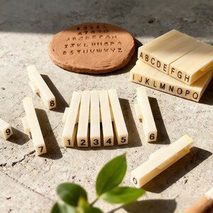 May include: A collection of cream-colored number and letter stamps, with a terracotta disc imprinted with numbers and the alphabet. The stamps are arranged on a light-colored surface, with a few green leaves in the foreground.
