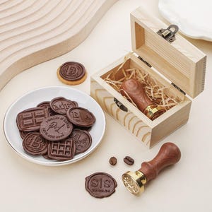 Puede incluir: Una caja de madera con tapa con bisagras, que contiene un sello de cera con mango de madera y base de metal dorado. Un plato blanco contiene varios sellos de cera marrón con varios diseños y textos en relieve. Sellos de cera y granos de café adicionales están dispersos.