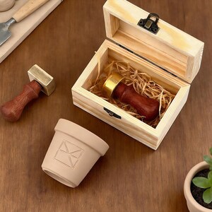 May include: A wooden box with a brass clasp holds a wax seal stamp with a dark wood handle and gold-colored stamp head. A second stamp rests beside the box. A small, beige pot with an envelope design is also present.