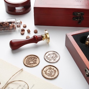 May include: A wax seal stamp with a dark wood handle and brass head, accompanied by a wooden storage box, a glass vial of small brown wax beads, and three circular wax seals featuring embossed floral designs and the letters U, J, and DJ.