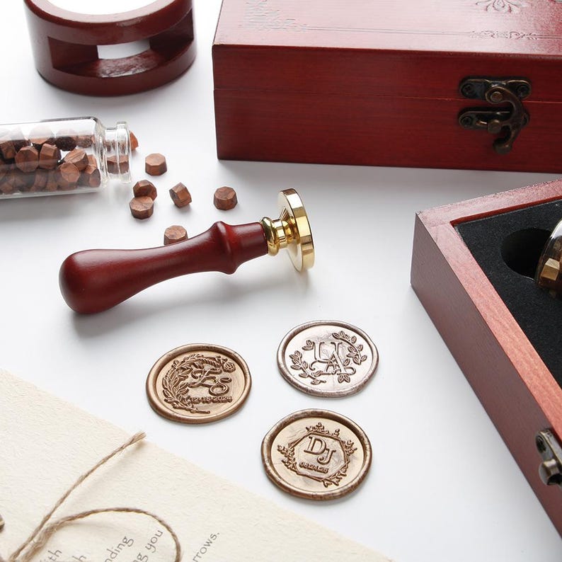 May include: A wax seal stamp with a dark wood handle and brass head, accompanied by a wooden storage box, a glass vial of small brown wax beads, and three circular wax seals featuring embossed floral designs and the letters U, J, and DJ.
