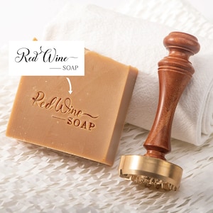 May include: A wooden stamp with a brass base and a red wine soap logo. The logo reads "Red Wine - SOAP". The stamp is used to imprint the logo onto a bar of soap.