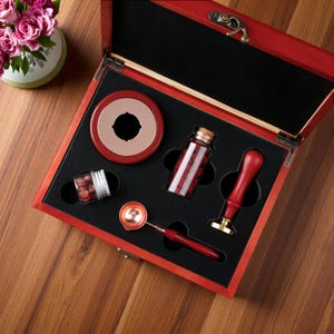 May include: A red wooden box containing a wax seal kit. The set includes a red wax seal stamp with a brass head, a metal melting spoon with a red handle, a round red wax warmer, and two glass jars filled with small red wax beads for sealing.