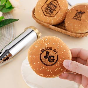 Puede incluir: Primer plano de panecillos de hamburguesa con semillas de sésamo. Una mano sostiene un panecillo con el texto "MADE BY JG" impreso. Otros panecillos están en una cesta de mimbre, con los textos "Sweet Island Burgers" y "ENRIQUE".
