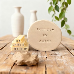 May include: A still life featuring pottery items. A round, beige pottery piece with the text "POTTERY BY CHRIS" is displayed next to a stack of rectangular blocks with the same text. Two white vases are in the background.