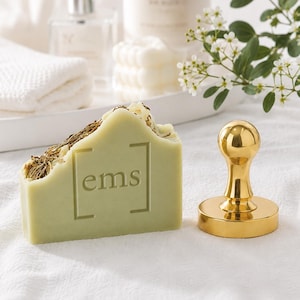 May include: A rectangular bar of light green soap topped with dried herbs and featuring the text ems enclosed in brackets, positioned next to a polished gold metal stamp with a rounded handle on a white textured fabric surface.