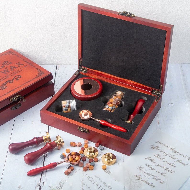May include: A dark wood storage box containing a wax seal kit with a metal melting spoon, two glass jars of wax beads, a wooden handle stamp, and a circular base. Outside the box are two additional stamps, a melting spoon, and wax beads.