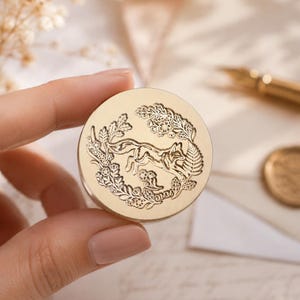 May include: A circular gold-toned wax seal stamp featuring an engraved design of a fox surrounded by a wreath of leaves and flowers. The stamp face displays intricate botanical details and a small fox figure in the center, crafted from metal.