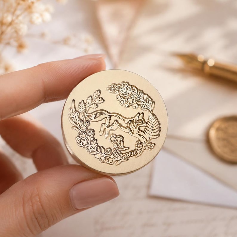 May include: A circular gold-toned wax seal stamp featuring an engraved design of a fox surrounded by a wreath of leaves and flowers. The stamp face displays intricate botanical details and a small fox figure in the center, crafted from metal.