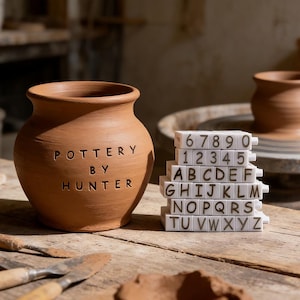 Puede incluir: Un jarrón de cerámica de terracota con la inscripción "POTTERY BY HUNTER" se muestra sobre una superficie de madera. Bloques blancos con letras y números están al lado. La imagen evoca un estudio de cerámica.