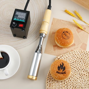 May include: A branding iron with a wooden handle and digital control panel, next to two sesame seed hamburger buns with branded designs. One bun says "MADE BY JLD" and the other says "RAKONICK" with a flame graphic.