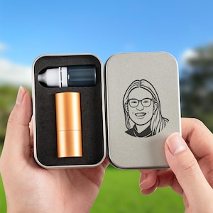 May include: A rectangular silver metal tin containing a small white-capped dropper bottle and a cylindrical gold-colored container set in black foam. The inside of the lid features a black line-art illustration of a person wearing glasses.