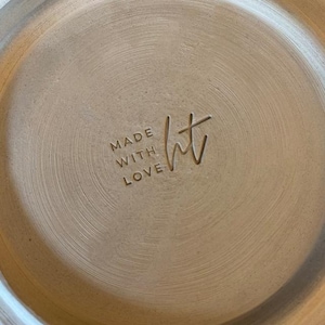 Puede incluir: Primer plano de un plato de cerámica beige con las palabras "MADE WITH LOVE" y las iniciales "ht" grabadas en la superficie. El plato tiene un diseño circular texturizado.