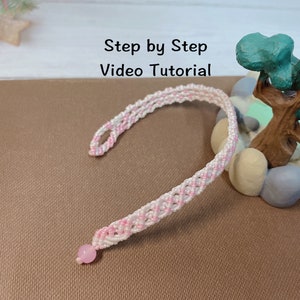 Può includere: Un braccialetto intrecciato rosa e bianco con un ciondolo rosa. Il braccialetto è su una superficie marrone con un piccolo albero e pietre sullo sfondo. Il testo "Step by Step Video Tutorial" è in alto nell'immagine.