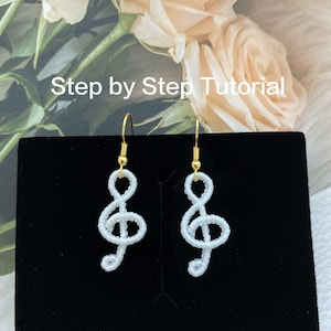 Op de afbeelding: Witte kralen oorbellen in de vorm van vioolsleutels, hangend aan gouden haken. De oorbellen worden getoond op een zwarte achtergrond. "Step by Step Tutorial" staat boven de oorbellen.