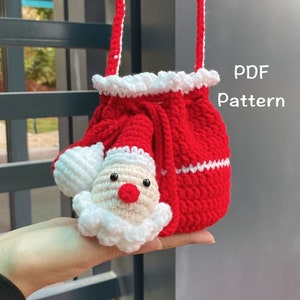 May include: A red and white crocheted Santa Claus drawstring bag with a white crocheted Santa Claus face on the front. The bag has a white crocheted trim around the top and a white crocheted strap. The text "PDF Pattern" is visible in the image.