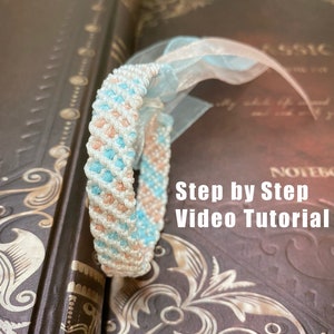 May include: A handmade macrame bracelet in white, light blue, and peach colors, with a light blue ribbon. The bracelet is resting on a vintage book. The text "Step by Step Video Tutorial" is visible.