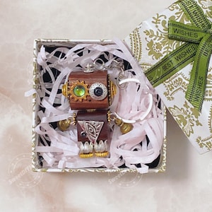 Puede incluir: Un llavero de robot de madera hecho a mano con detalles en latón y plata, presentado en una caja de regalo decorativa con papel triturado. El robot presenta detalles en forma de engranaje y ojo. La caja de regalo está envuelta con una cinta verde que dice "Best Wishes".