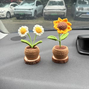 May include: Two crocheted potted plants sitting on a car dashboard. One pot contains two white daisies with yellow centers and green stems. The other pot holds a single large sunflower with yellow petals, a brown center, and green leaves.