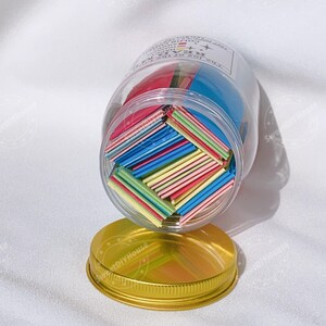Op de afbeelding: Een glazen pot gevuld met kleurrijke papieren vierkanten. De pot heeft een gouden deksel en een label met de tekst "Deze set van 100+ DIY KARAOKE PAPIER is voor jou om van te genieten!"
