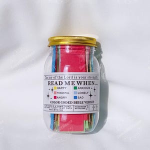 Op de afbeelding: Een glazen pot met een gouden deksel gevuld met kleurrijke papiertjes. De pot heeft een label met de tekst "Lees me wanneer..." met een lijst van emoties en bijbehorende kleuren. Het label zegt ook "Kleurgecodeerde Bijbelverzen".