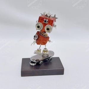 以下が含まれることがあります： 木と金属で作られた手作りのロボットの彫刻。ロボットはスケートボードに乗っています。ロボットは赤い木の体、大きな宝石の目、王冠を持っています。スケートボードは銀色で、ベースは濃い茶色です。