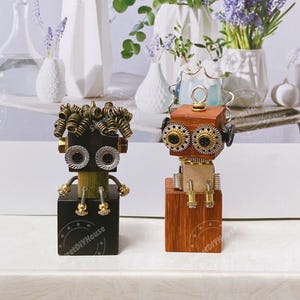 May include: Two small, handmade robot figurines made of wood and metal. The robot on the left has a black body, gold and silver accents, and a curly hairpiece made of metal springs. The robot on the right has a brown wooden body, gold and silver accents, and a metal crown. Both robots are standing on wooden bases.