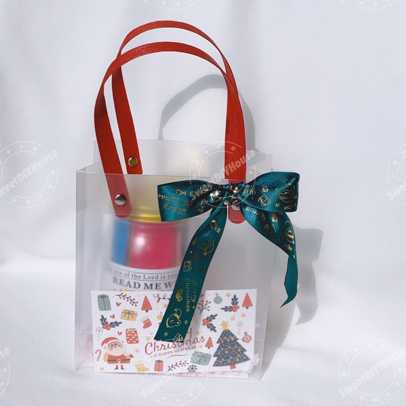 Op de afbeelding: Een transparante plastic cadeauzak met rode handvatten en een groen lint met een strik. De tas bevat een klein cadeaudoosje met een rood en blauw ontwerp en een kaart met de tekst "Kerstmis & Gelukkig Nieuwjaar".