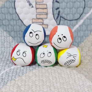 May include: Five colorful, round plush toys with expressive cartoon faces. Each ball has a white center with a different colored top and bottom, including red, blue, green, and yellow. The faces display various emotions.