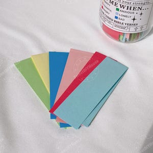 Op de afbeelding: Een set van zes kleurrijke papieren stroken met de tekst "ME WHEN..." en een lijst met emoties en bijbehorende kleuren. De tekst "COLOR CODED BIBLE VERSES" is ook zichtbaar.