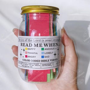 Op de afbeelding: Een glazen pot gevuld met gekleurde papieren stroken. De pot heeft een gouden deksel en een wit etiket met de tekst "Read Me When..." met een lijst van emoties en bijbehorende kleuren. Het etiket vermeldt ook "Color Coded Bible Verses" en "Handmade with Love".