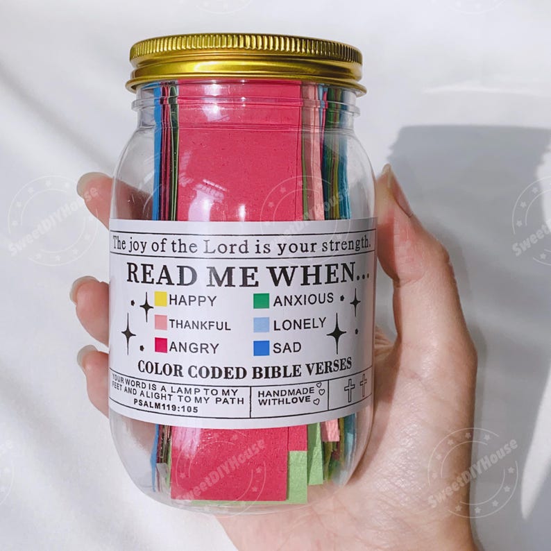 Op de afbeelding: Een glazen pot gevuld met gekleurde papieren stroken. De pot heeft een gouden deksel en een wit etiket met de tekst "Read Me When..." met een lijst van emoties en bijbehorende kleuren. Het etiket vermeldt ook "Color Coded Bible Verses" en "Handmade with Love".