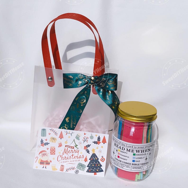 Op de afbeelding: Een transparante plastic cadeauzak met rode handvatten en een groen-gouden lint. De zak bevat roze tissuepapier en een kerstkaart. Naast de zak staat een glazen pot met een gouden deksel gevuld met gekleurde papieren stroken. De pot heeft een label met de tekst "Lees me wanneer" en somt verschillende emoties op.