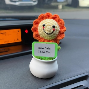 Pode incluir: Uma flor de crochê com pétalas laranja e um rosto cor de creme com olhos pretos, um sorriso bordado e bochechas rosadas. A flor está num pequeno vaso branco com uma placa verde que diz em texto preto: "Drive Safe I Love You".