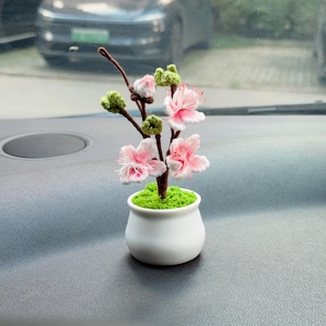May include: A small crochet cherry blossom tree in a white ceramic pot. The tree features brown branches, green buds, and light pink flowers with white centres. The base of the tree is covered in bright green moss-like material inside the pot.