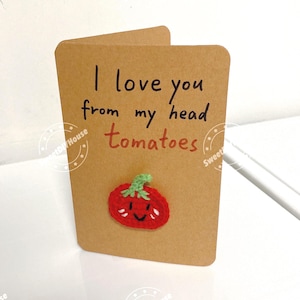 May include: A tan greeting card featuring the handwritten black text I love you from my head and the red text tomatoes, with a small, round, red crocheted tomato applique attached to the front, complete with a green stem and a smiling face.