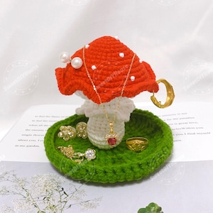 Peut inclure: Un porte-bijoux en forme de champignon crocheté. Le champignon a un chapeau rouge à pois blancs, un pied blanc et une base verte. Des bijoux en or, dont des boucles d'oreilles, des bagues et des colliers, sont exposés sur le support. Le porte-bijoux est posé sur un livre.