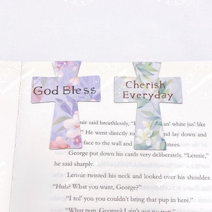Könnte beinhalten: Zwei kreuzförmige Lesezeichen mit floralen Mustern. Eines trägt die Aufschrift "God Bless", das andere "Cherish Everyday". Die Lesezeichen sind hellviolett mit rosa und weißen floralen Akzenten und liegen auf einer Buchseite.