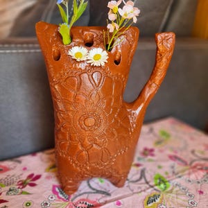 Könnte beinhalten: Eine handgefertigte, braune Keramikvase mit einem einzigartigen katzenartigen Design. Die Vase weist ein detailliertes Blumenmuster auf und enthält kleine Blumen mit weißen, rosa und blauen Blütenblättern. Sie hat einen Henkel und drei kleine Füße.