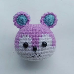 May include: A crocheted stuffed animal with a purple body, white face, and blue ears. The animal has black eyes and a stitched smile.