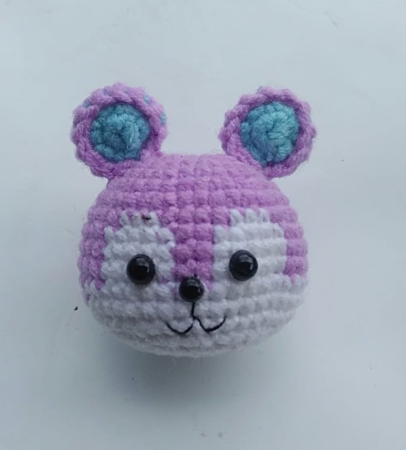 May include: A crocheted stuffed animal with a purple body, white face, and blue ears. The animal has black eyes and a stitched smile.