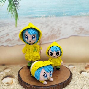 Könnte beinhalten: Drei gehäkelte Puppen, die gelbe Regenmäntel und blaues Haar tragen. Die Puppen stehen auf einer Holzplattform. Die größte Puppe hält einen Stock. Die kleinste Puppe liegt auf dem Bauch.
