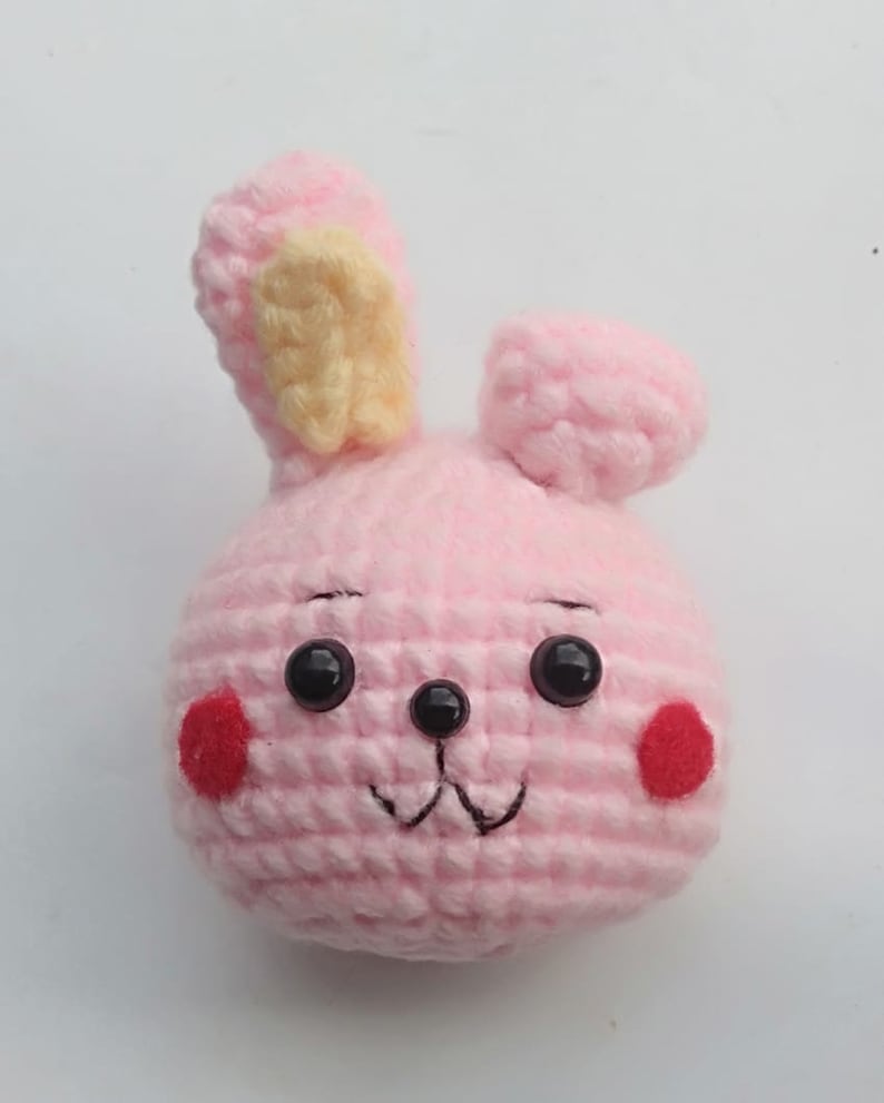May include: Pink crocheted bunny with black eyes, a black nose, and red cheeks. The bunny has a yellow crocheted ear.