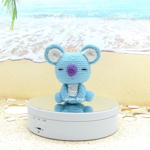 K&ouml;nnte beinhalten: Ein hellblaues geh&auml;keltes Koala-Pl&uuml;schtier mit wei&szlig;en Ohren und einer lila Nase. Das Spielzeug sitzt auf einem wei&szlig;en drehbaren Pr&auml;sentationsst&auml;nder mit einer verspiegelten Oberfl&auml;che. Der St&auml;nder steht auf einem sandigen Strandhintergrund.