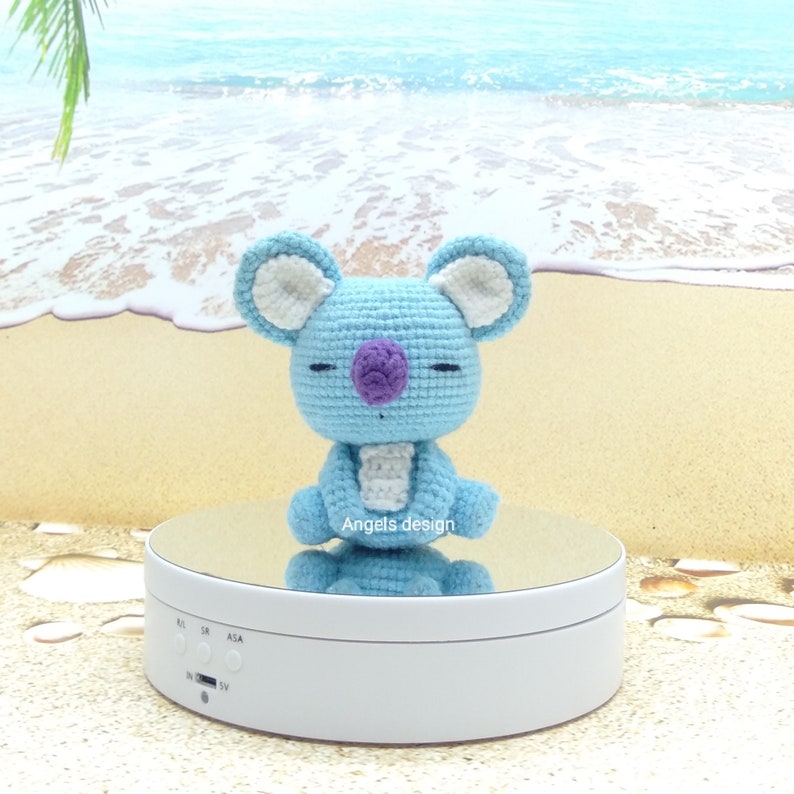 K&ouml;nnte beinhalten: Ein hellblaues geh&auml;keltes Koala-Pl&uuml;schtier mit wei&szlig;en Ohren und einer lila Nase. Das Spielzeug sitzt auf einem wei&szlig;en drehbaren Pr&auml;sentationsst&auml;nder mit einer verspiegelten Oberfl&auml;che. Der St&auml;nder steht auf einem sandigen Strandhintergrund.