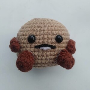May include: A crocheted plush toy with a brown body, brown arms, and black eyes. The toy has a white stitched mouth.