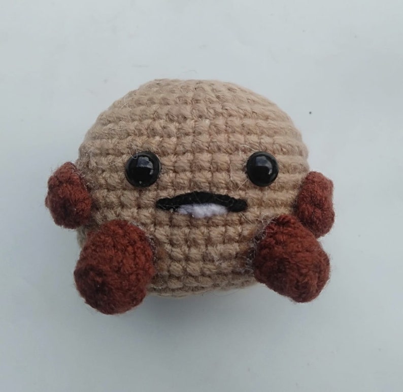 May include: A crocheted plush toy with a brown body, brown arms, and black eyes. The toy has a white stitched mouth.